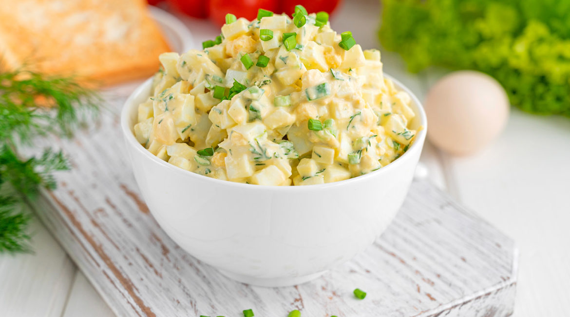 Egg salad with chopped green onions on top in a white bowl for cooking a sandwich on a white wooden background. Selective focus.